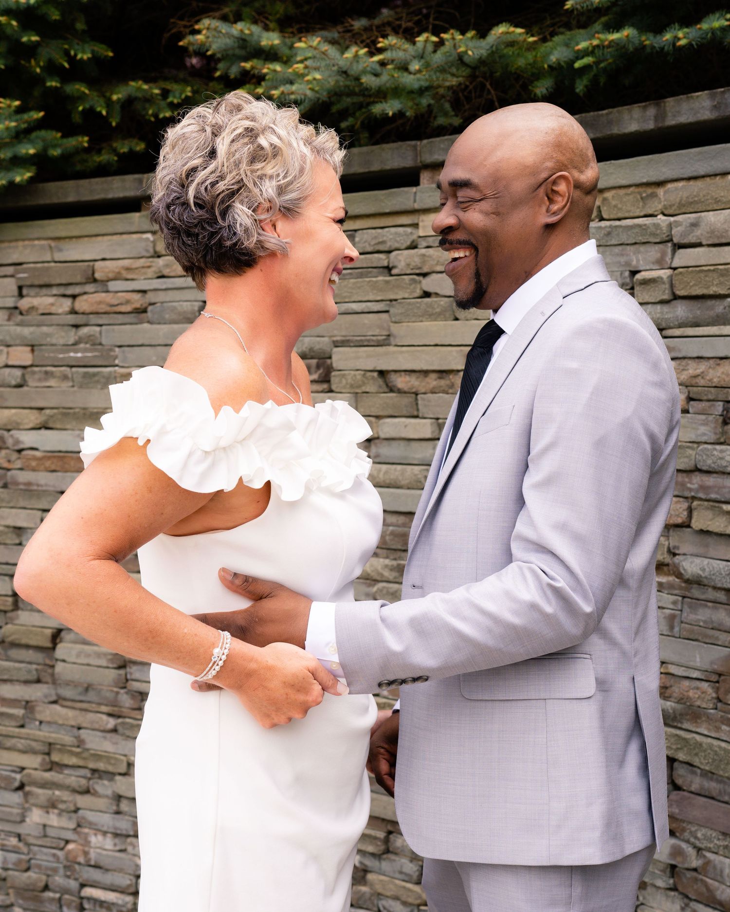 A joyful couple in wedding attire laughing together outdoors, bride in white off-shoulder gown, groom in light gray suit.