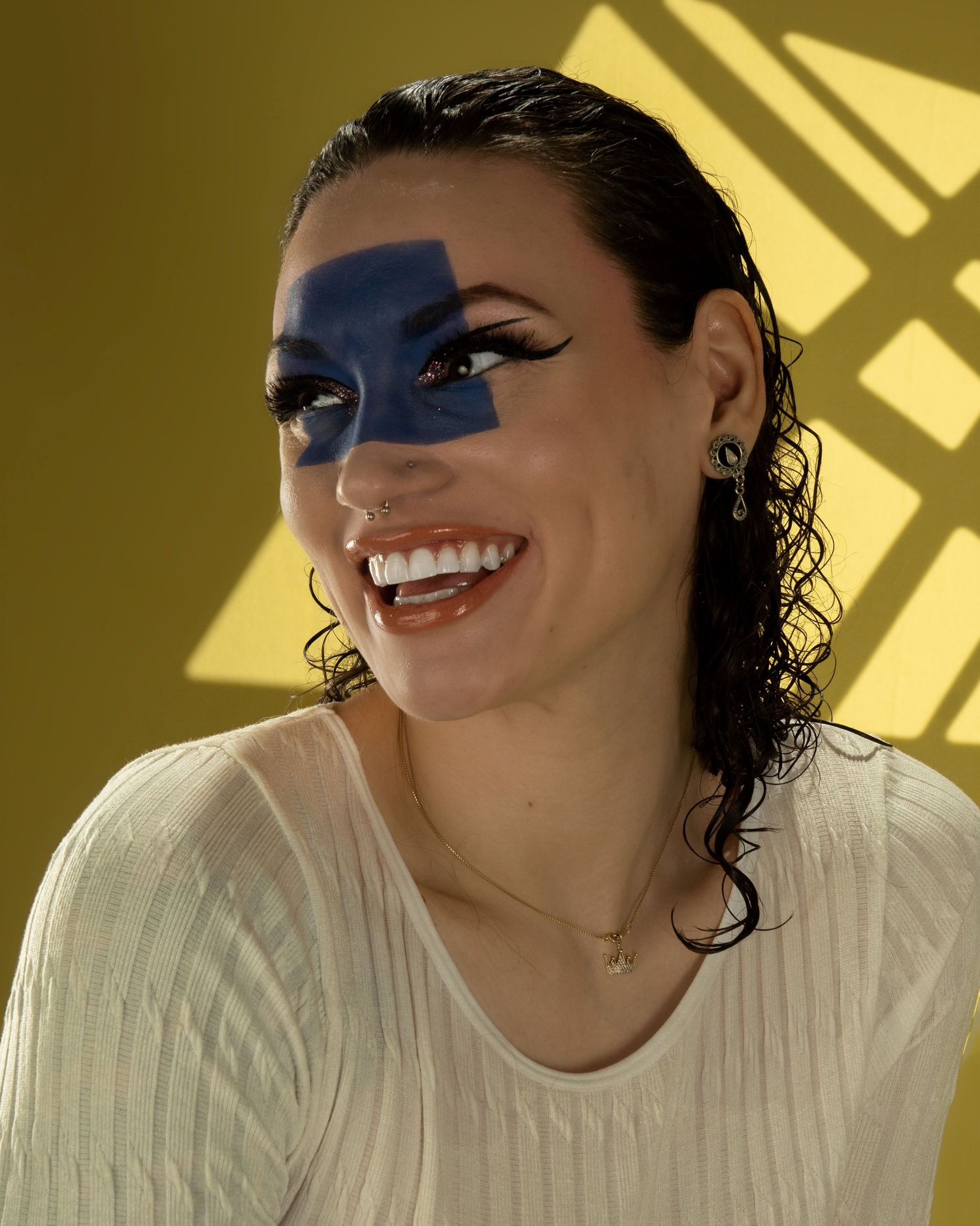 Smiling woman with blue geometric face paint, wet hair, wearing white top against yellow background.