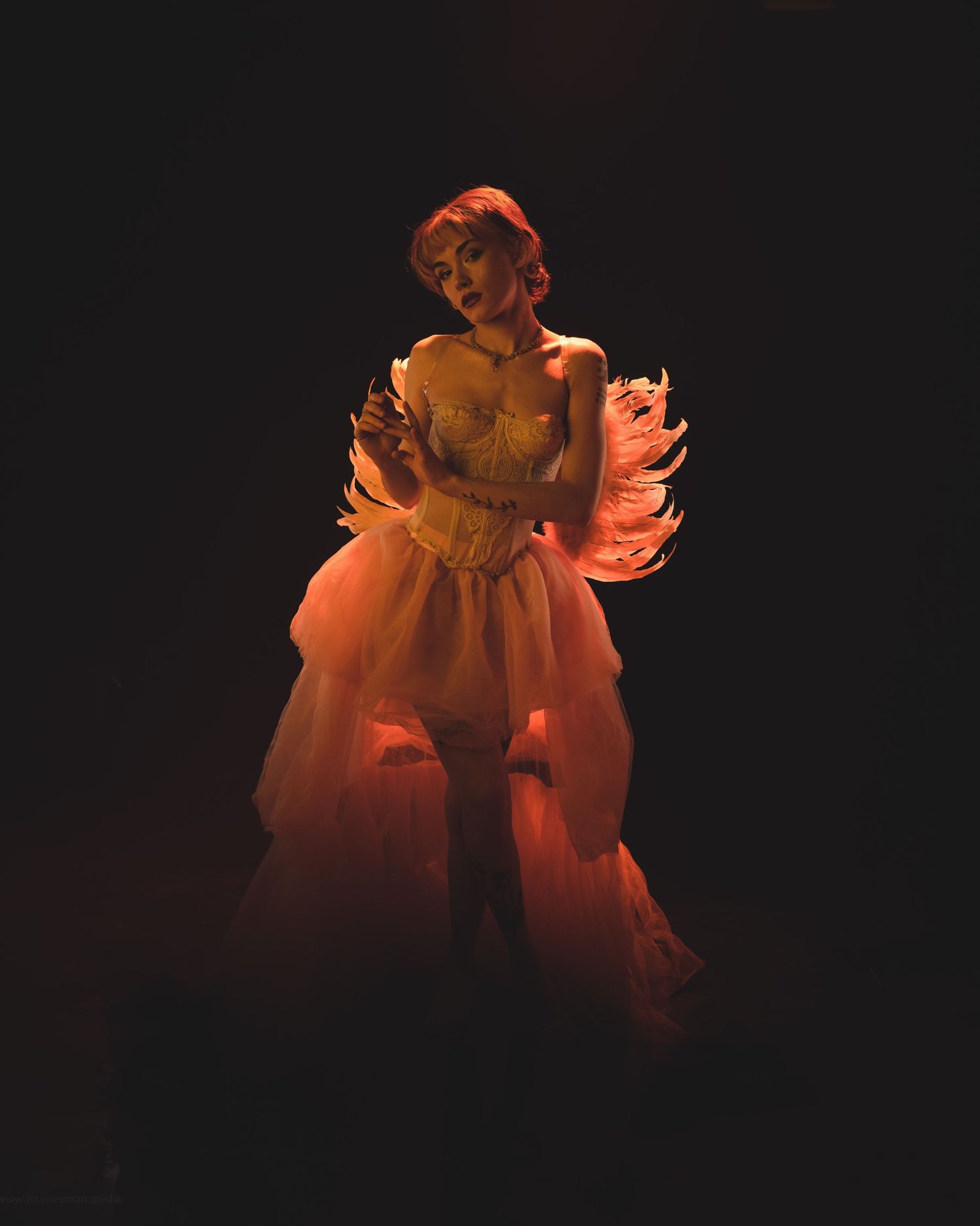 A performer in an ornate pink feathered corset gown poses dramatically under warm stage lighting against a dark background.