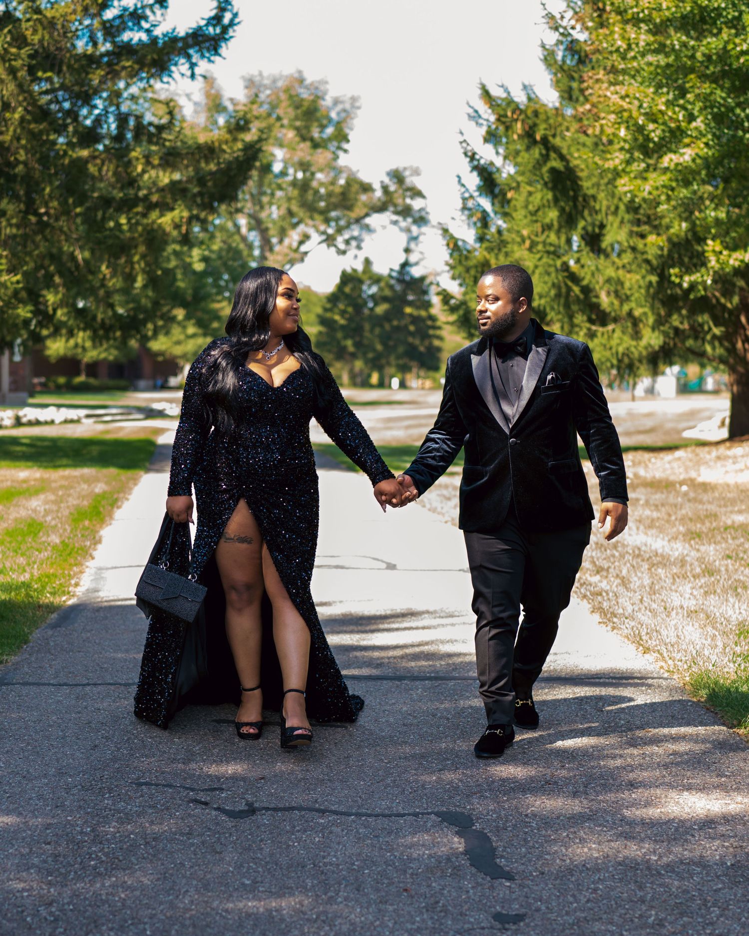 Elegant couple in matching black formal attire holding hands while walking along a tree-lined path outdoors.
