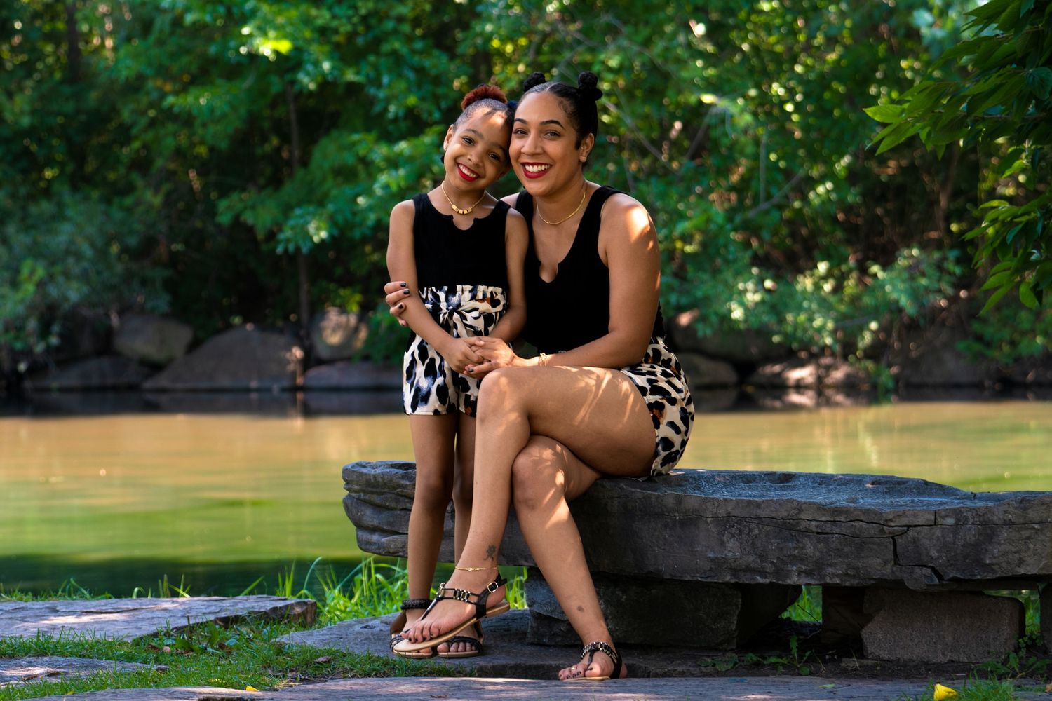 Two women sitting together on a stone ledge in a lush green park near a river, wearing stylish outfits.