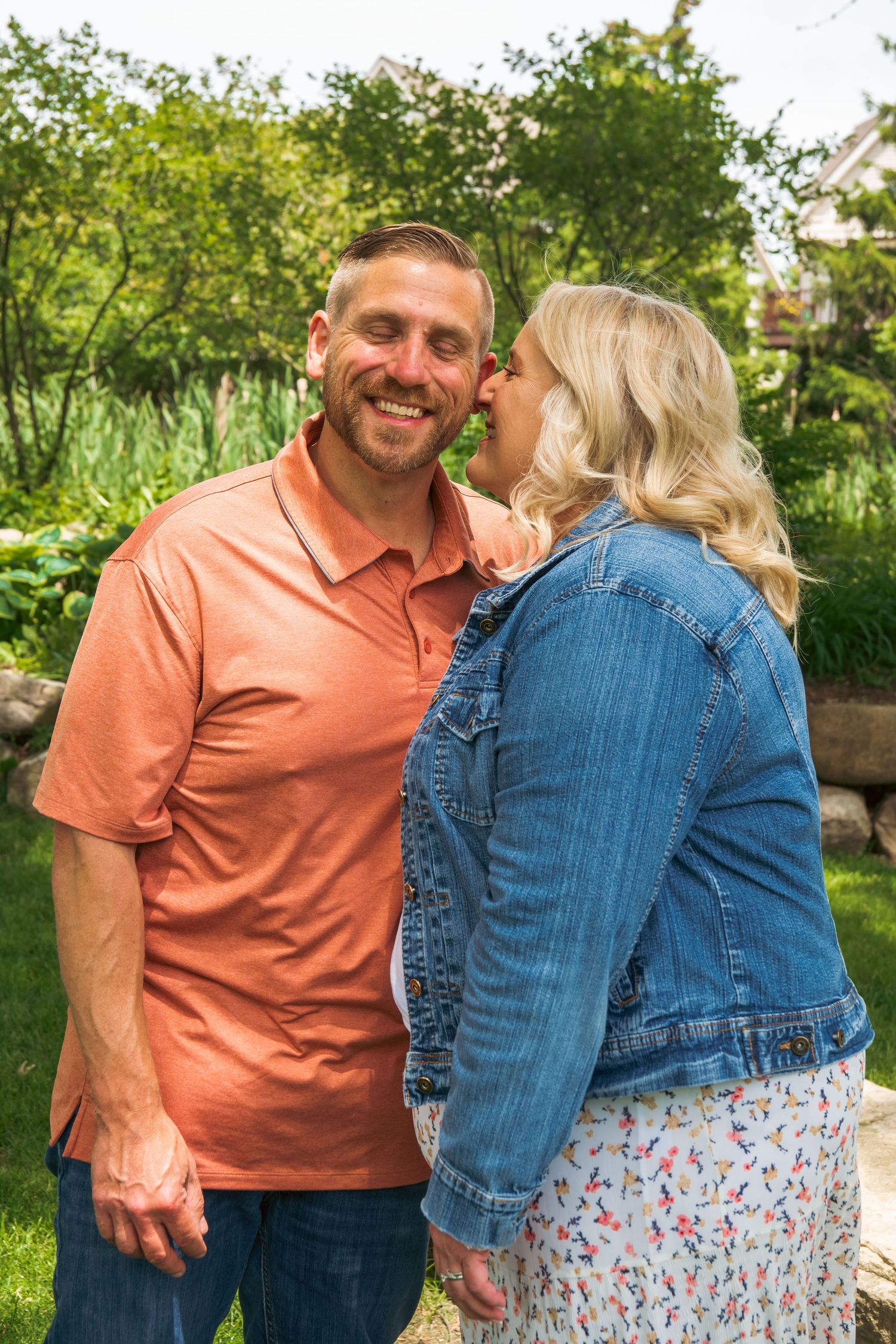 A couple in a denim jacket and coral shirt embrace outdoors in a green garden setting during a sunny day.