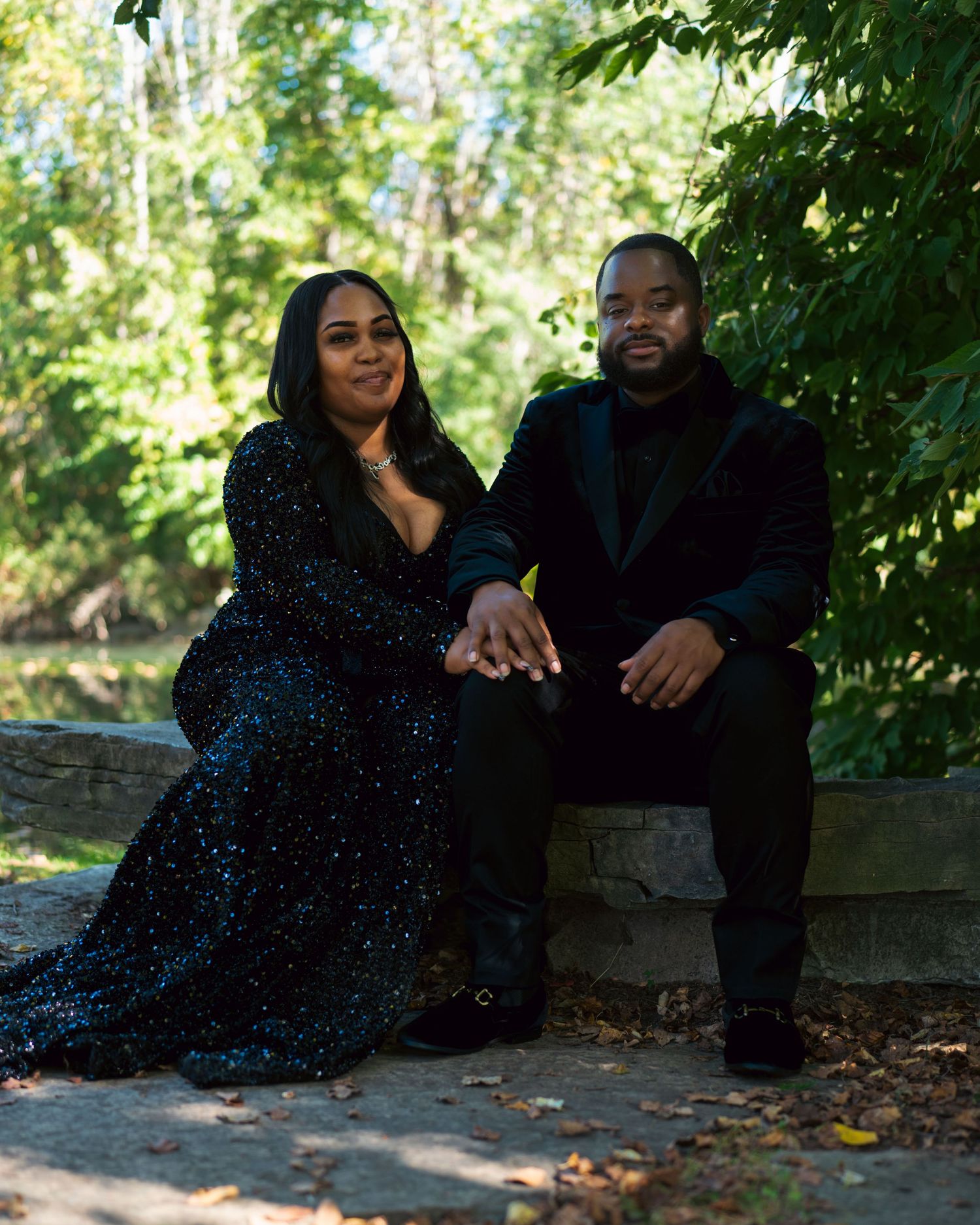 A couple sits together on a stone wall surrounded by lush greenery on a sunny autumn day.