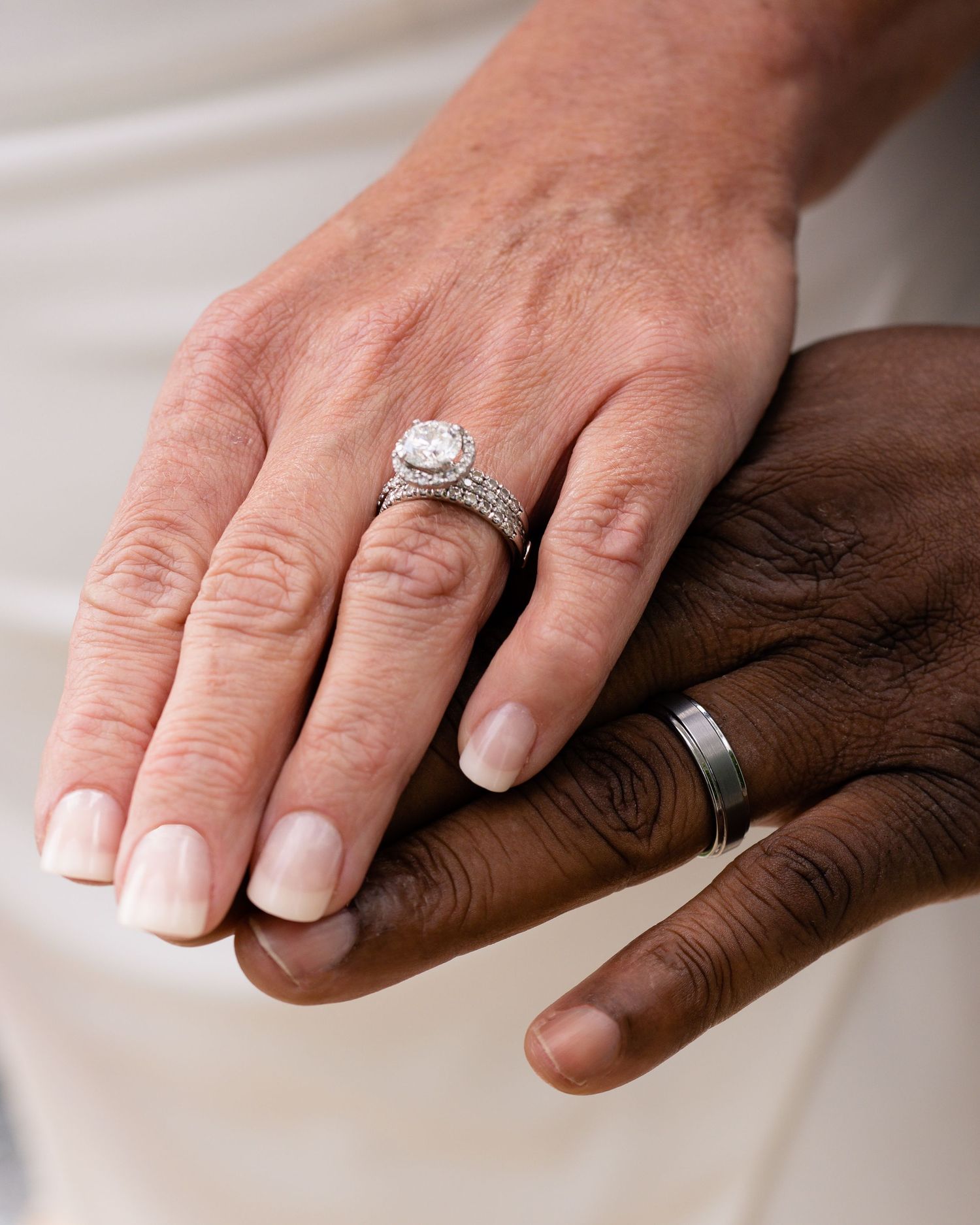 Two hands with wedding rings, one light-skinned with diamond halo ring, one dark-skinned with silver band, symbolizing marriage.