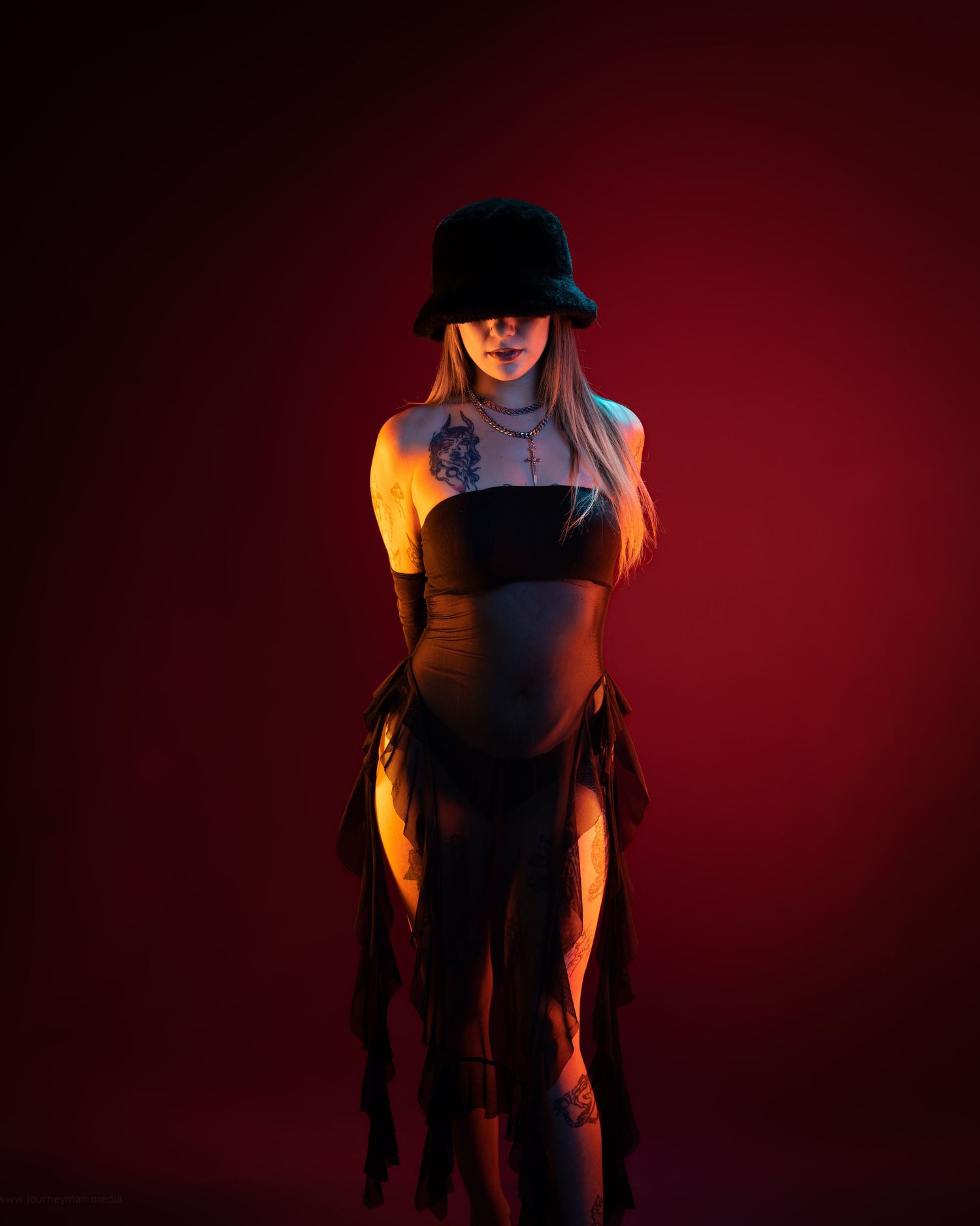 Woman in black bucket hat and strapless outfit with tattoos poses against a dramatic red-lit studio background.