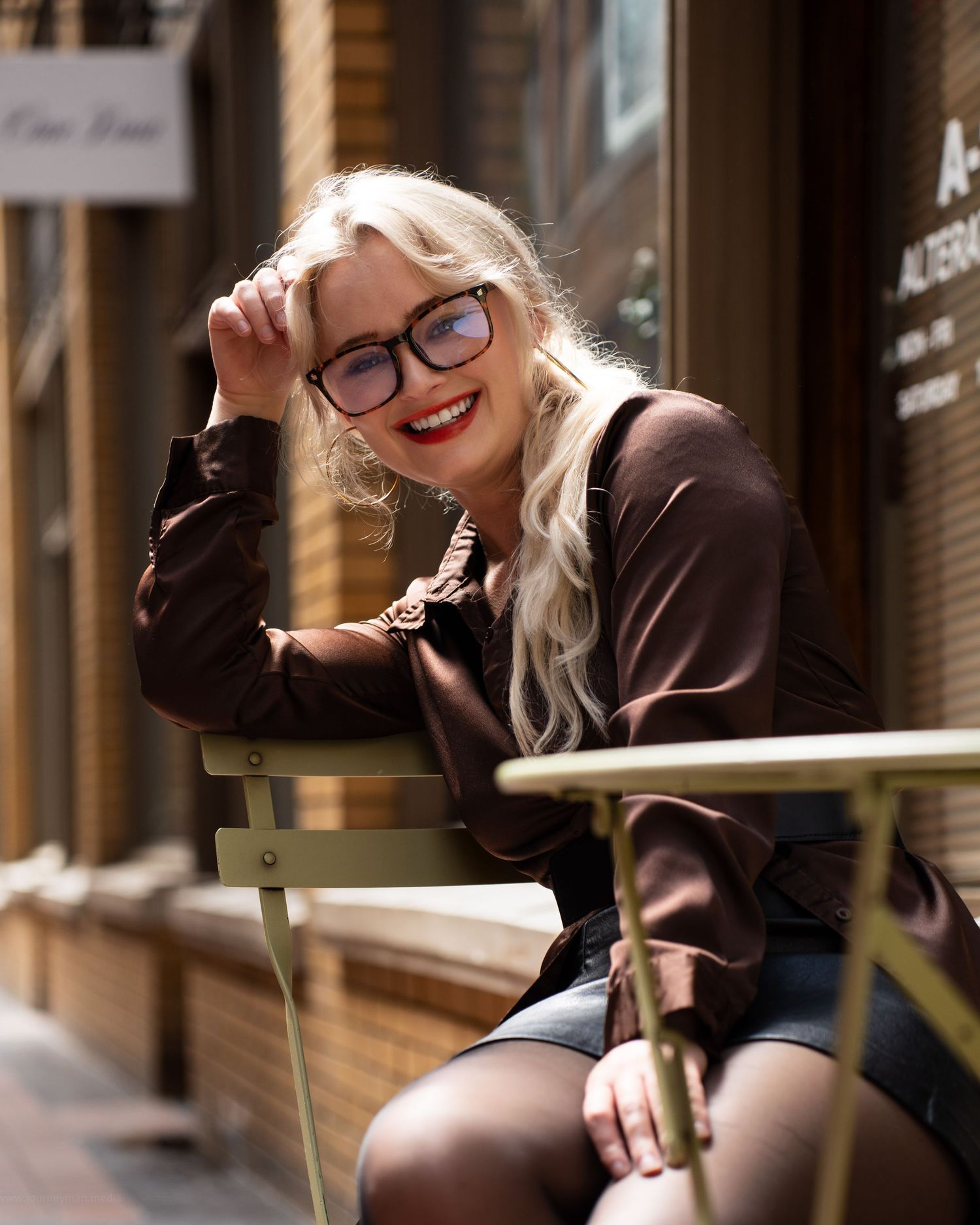 Smiling blonde woman with glasses in brown blouse sits at outdoor cafe table, leaning on hand.