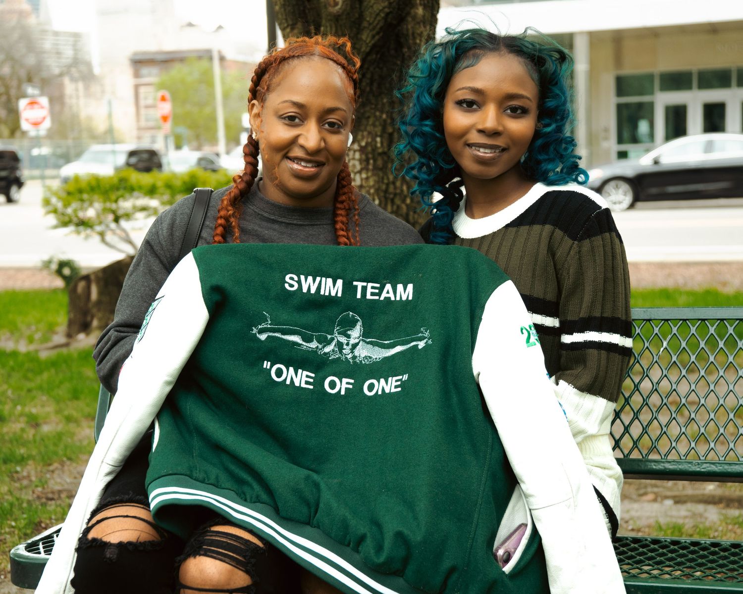 Two women holding a green Swim Team One of One shirt while sitting on a bench outdoors.