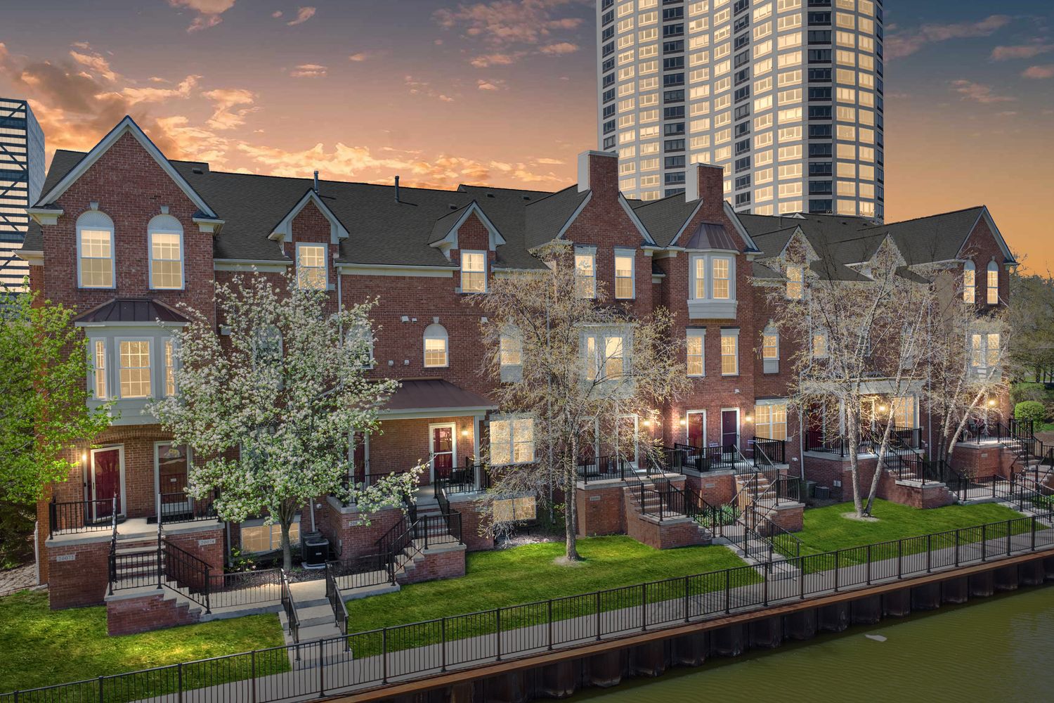 Row of townhouses with brick facades against dramatic sunset sky with high-rise buildings in background.