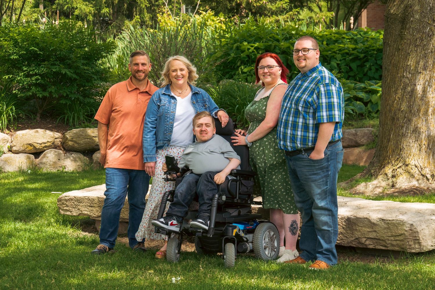 Group of five people smiling outdoors, one seated in a power wheelchair, posing together on green grass.
