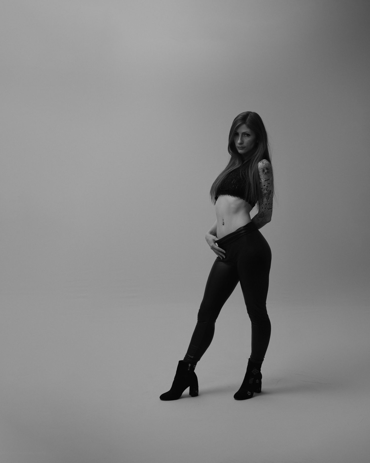 Black and white studio photo of a tattooed woman in crop top and leggings posing confidently in heeled boots.