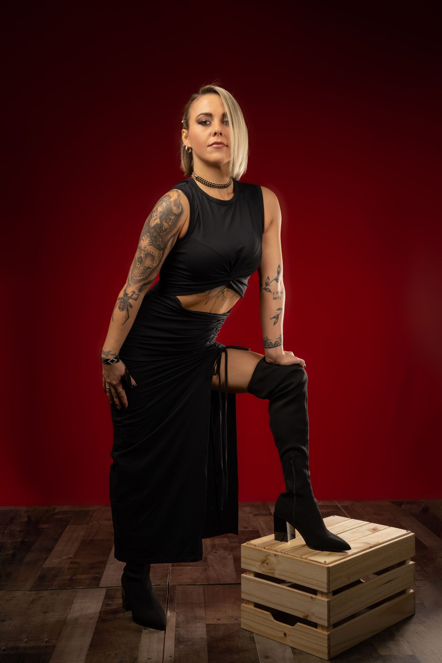 Person in black dress poses dramatically on wooden crates against dark red backdrop in artistic photoshoot series.