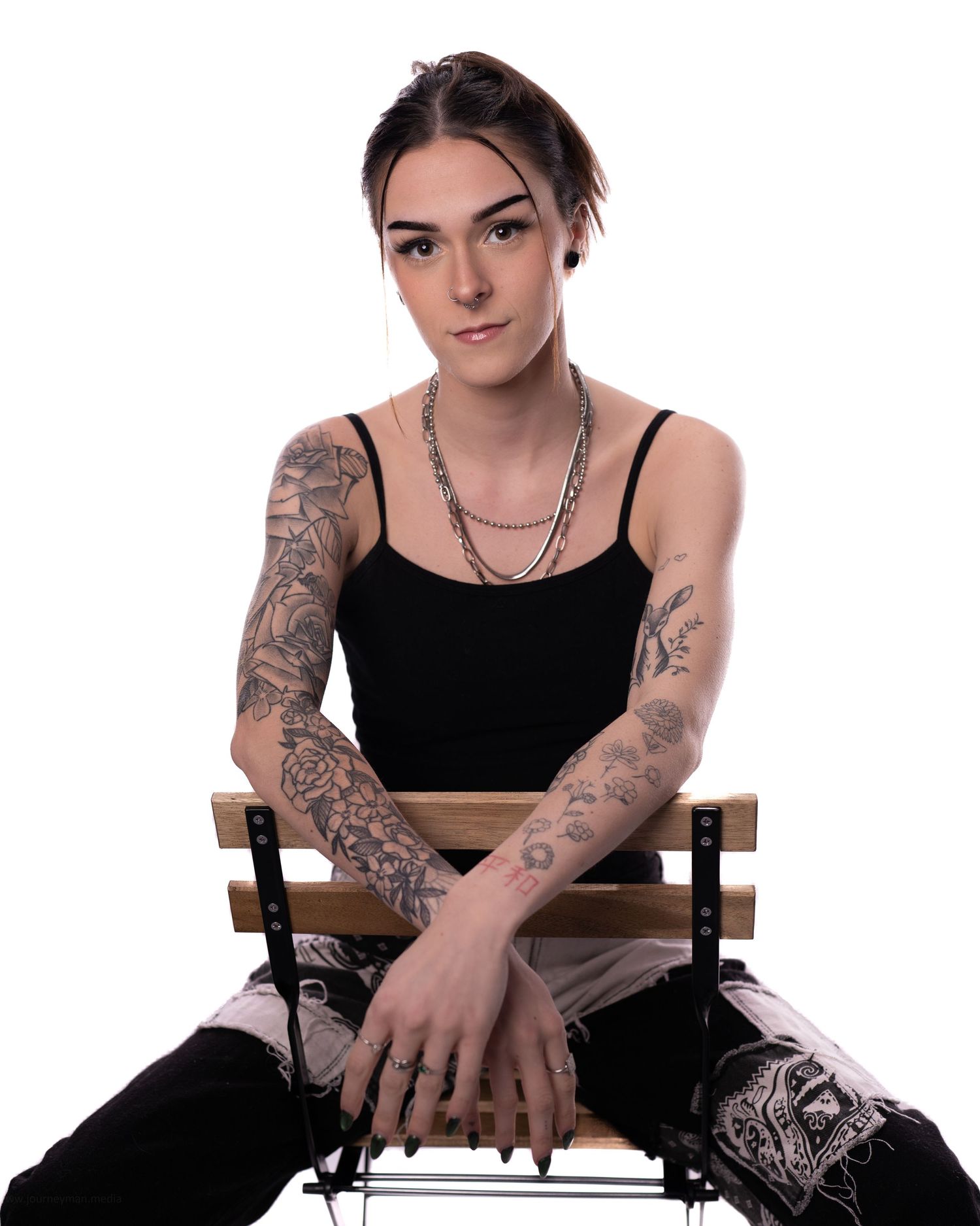 Tattooed young woman in black tank top sitting on wooden chair with arms crossed, wearing silver chain necklaces.