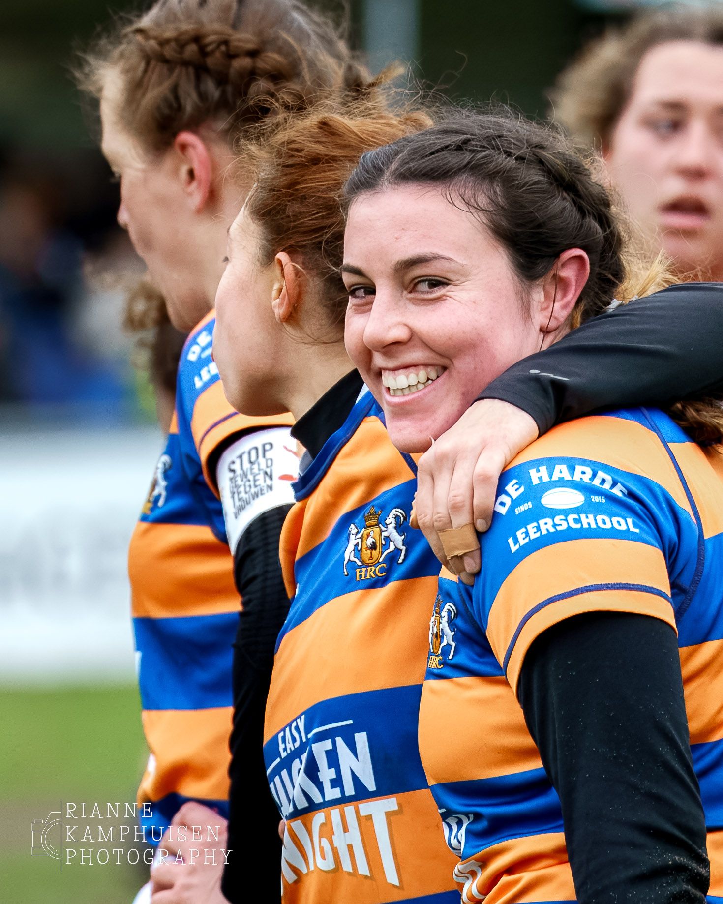 Vrouwelijke rugby speler lacht over haar schouder heen naar de camera. Ze heeft een oranje blauwe tenue aan. Regio Haaglanden