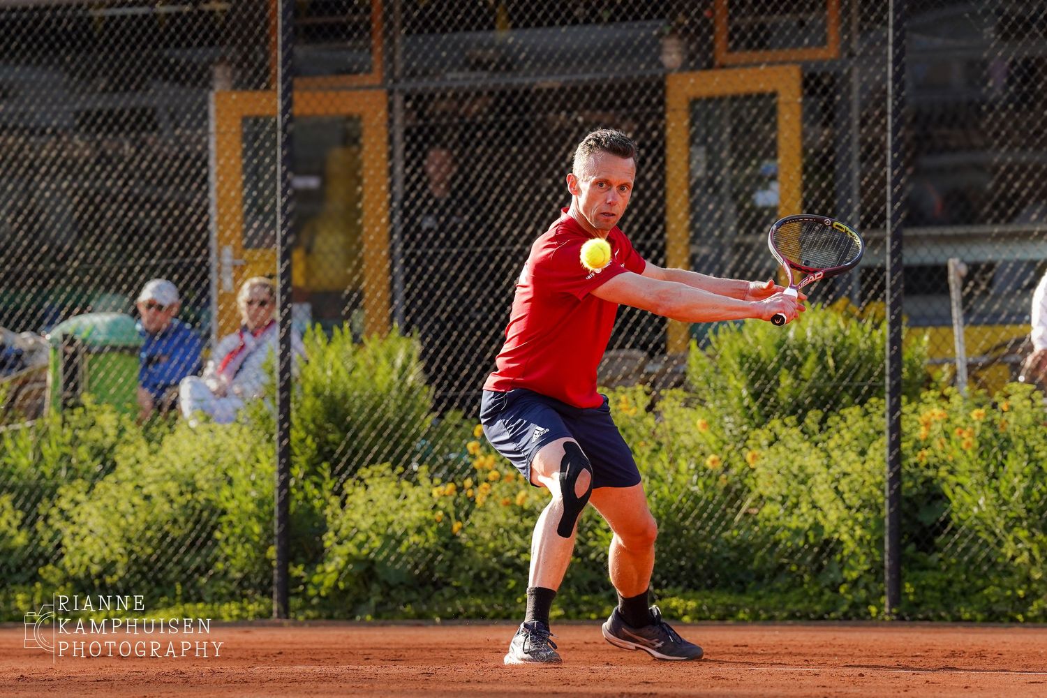Tennisser in rood shirt maakt een backhand-slag op het gravelveld, met focus op de bal.
