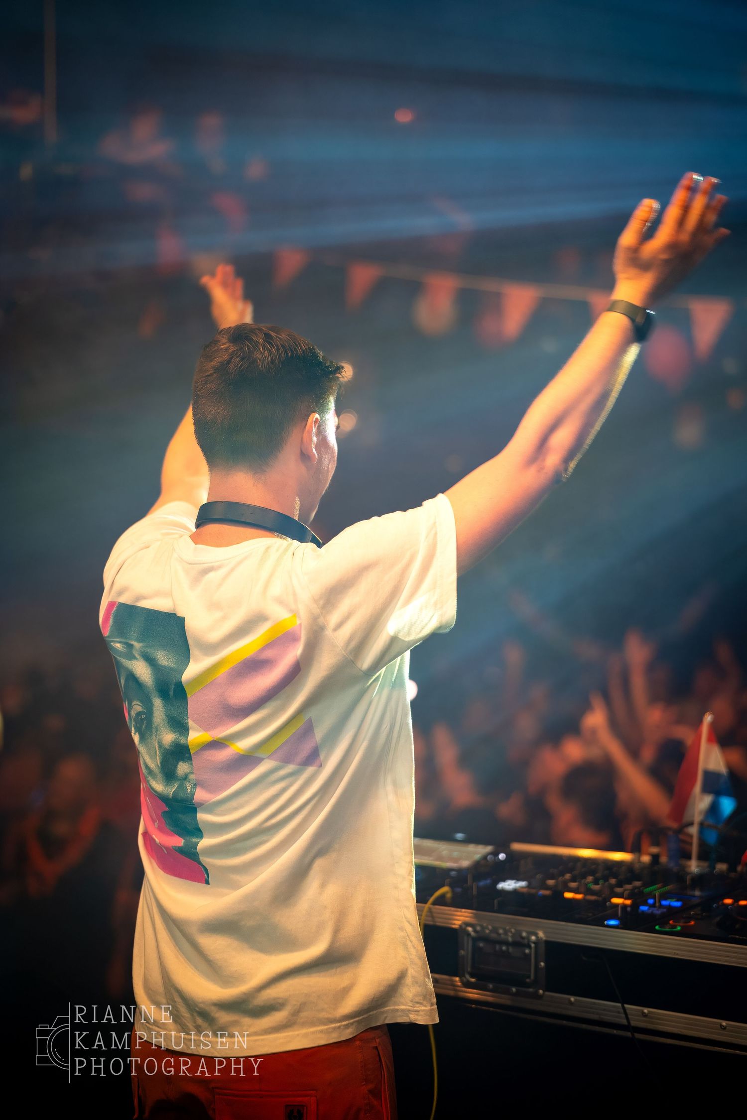 DJ met handen in de lucht kijkt uit over een uitzinnig feestpubliek tijdens een optreden vol licht en energie.