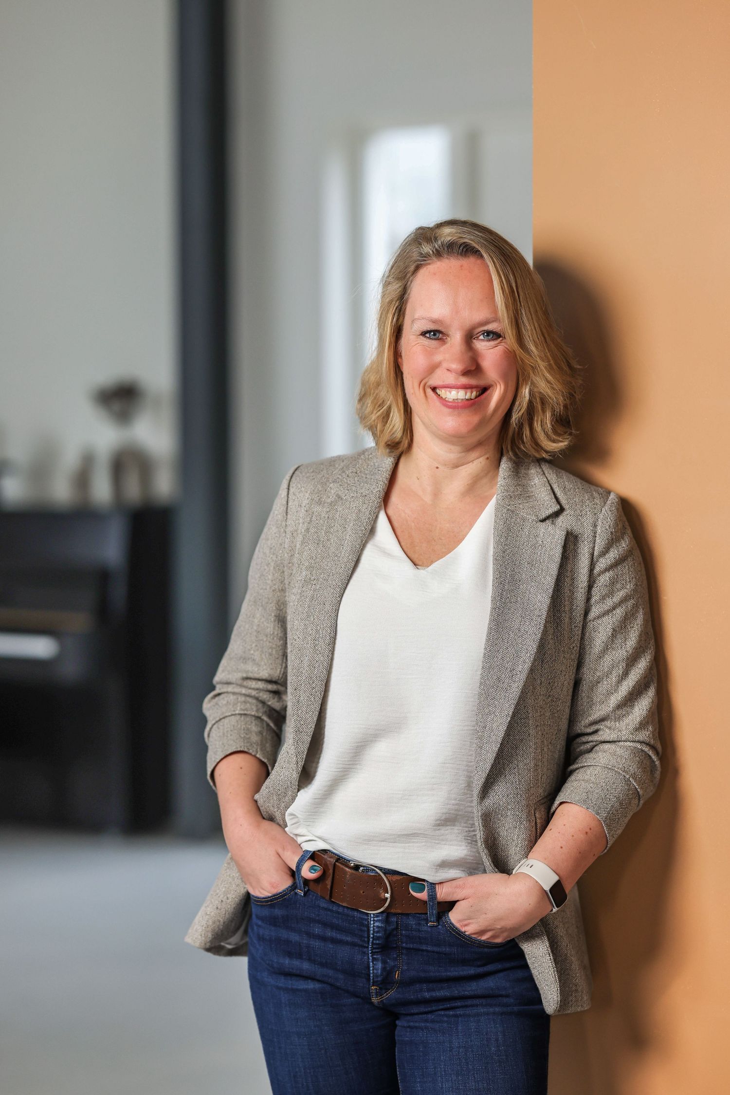 Zakelijke portretfoto van een vrouw die glimlachend tegen een oranje muur leunt, gekleed in blazer en jeans.