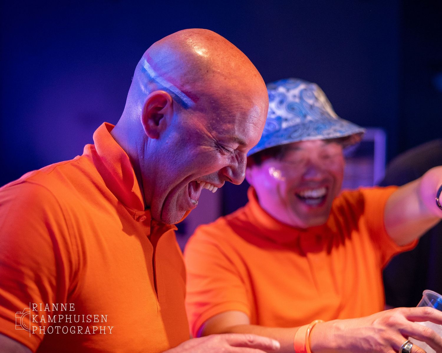 Twee mannen in oranje shirts lachen uitbundig achter een bar of DJ-tafel tijdens een feestelijke avond.