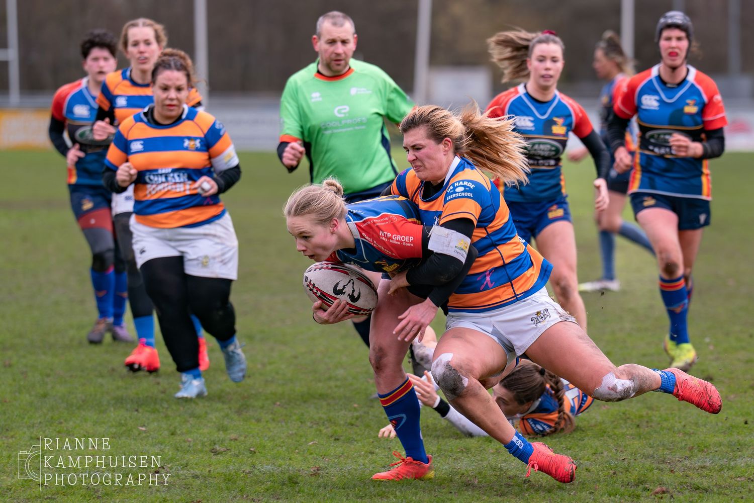 Damesrugby wedstrijd. Een dame tackelt tegenstander die rugbybal vast heeft. Op de achtergrond zie je andere spelers en scheidsrechter