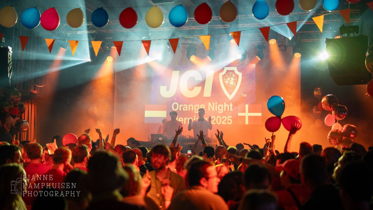 Drukke feestzaal tijdens JCI Orange Night 2025, met dansend publiek, oranje sfeerlicht en kleurrijke vlaggetjes en ballonnen boven podium