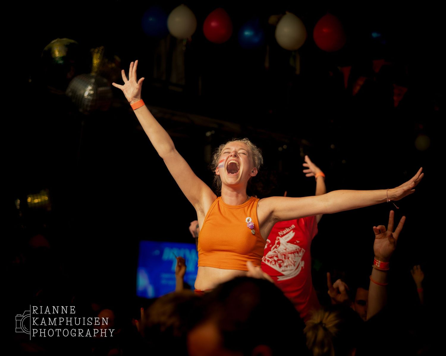 Vrouw in oranje top staat juichend op de schouders van iemand in het publiek tijdens een uitbundig feest.