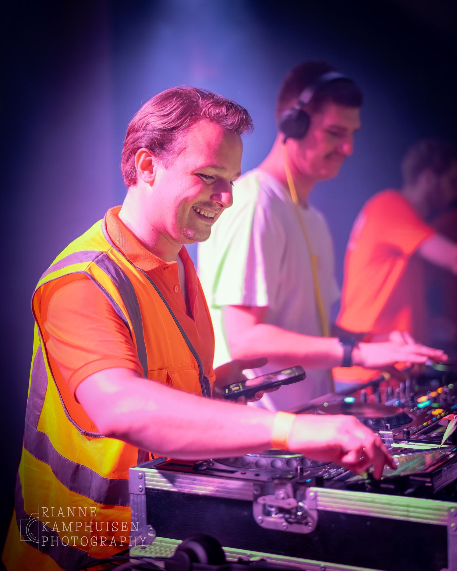DJ in oranje shirt en veiligheidshesje draait lachend muziek, naast twee collega-DJ’s in een kleurrijke setting.