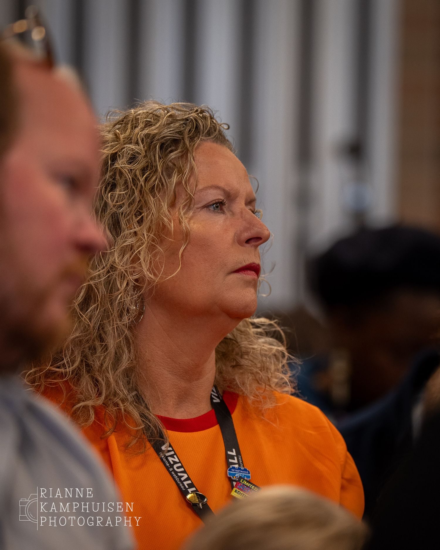 Vrouw in oranje shirt luistert geconcentreerd tijdens een bijeenkomst, met badge om haar nek.