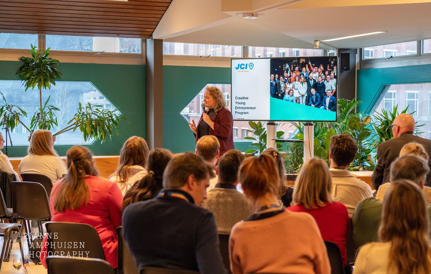 Spreker geeft presentatie voor een groep mensen tijdens een evenement, met een scherm op de achtergrond waarop 'JCI' en een groepsfoto te zi