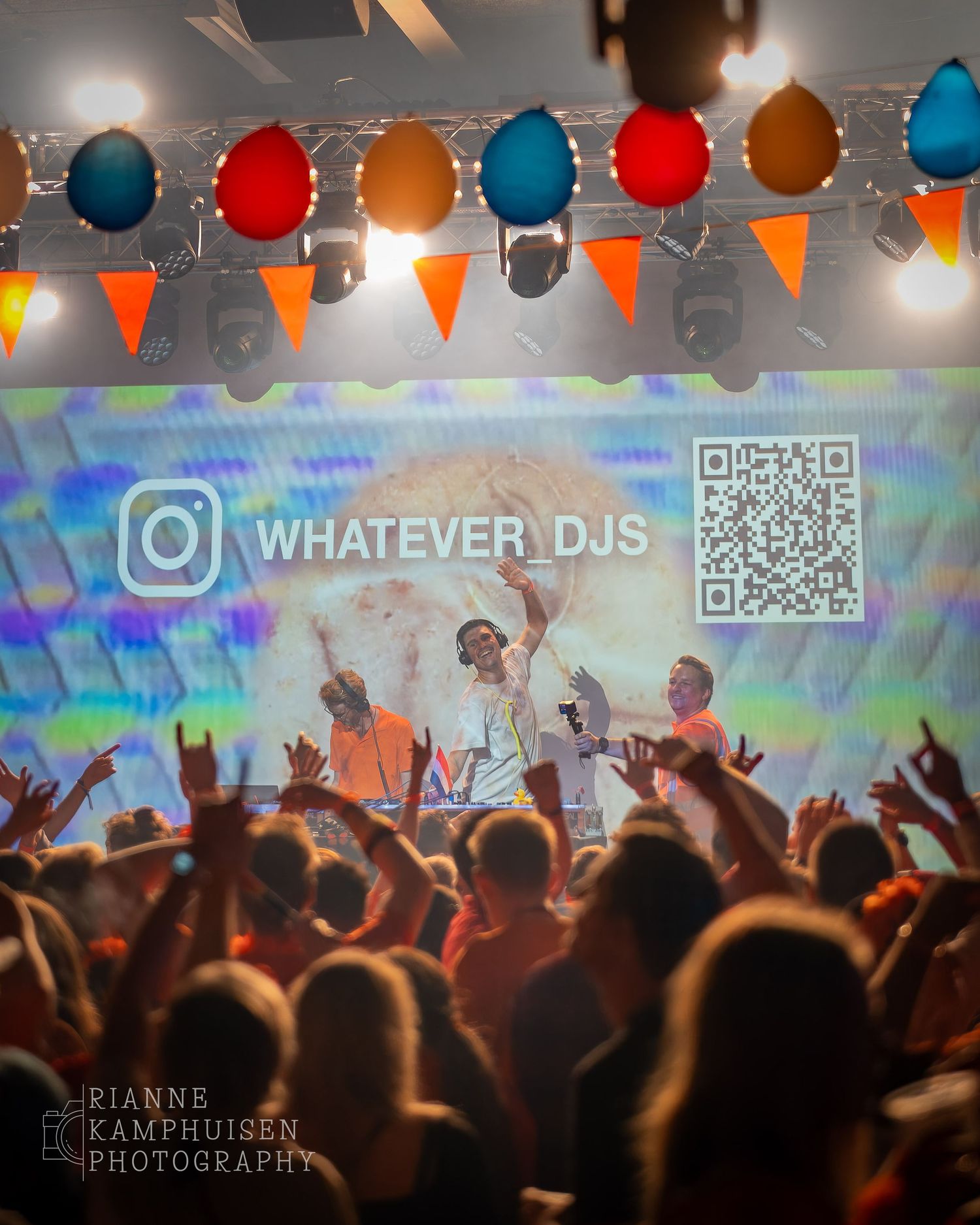 Publiek juicht richting het podium waar drie DJ’s onder kleurrijke vlaggetjes optreden, met projectie van “WHATEVER DJS” op het scherm.
