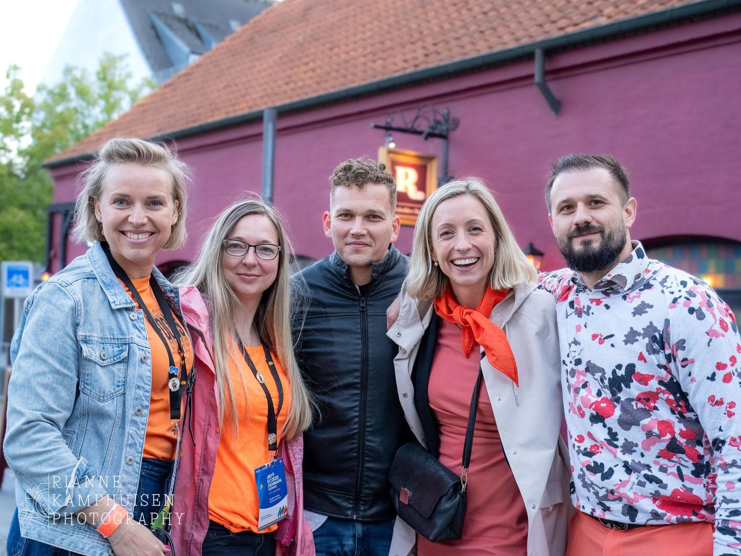 Groep van vijf mensen poseert lachend samen voor een rood gebouw tijdens een evenement in de avond.