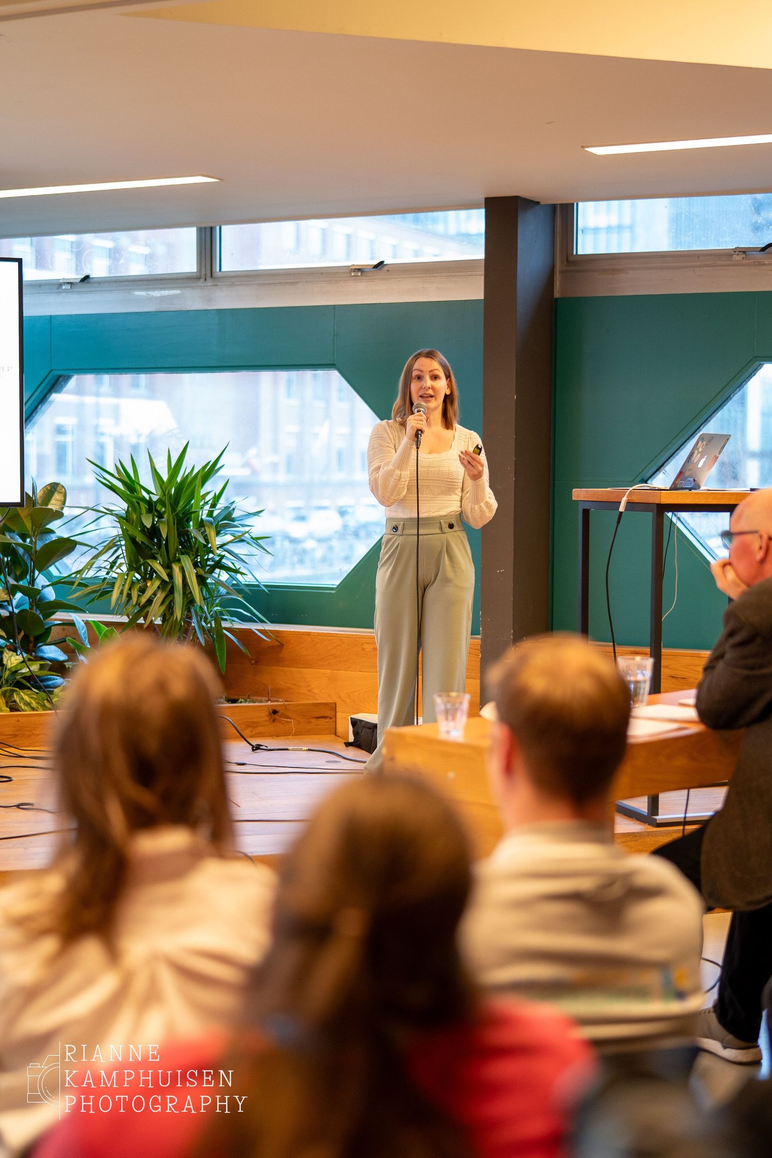 Vrouwelijke spreker staat op een podium met microfoon tijdens een evenement, terwijl het publiek aandachtig luistert.