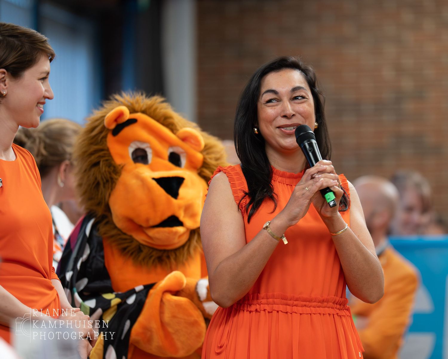 Vrouw in oranje jurk spreekt met microfoon, terwijl een mascotte in leeuwenpak vrolijk achter haar staat.