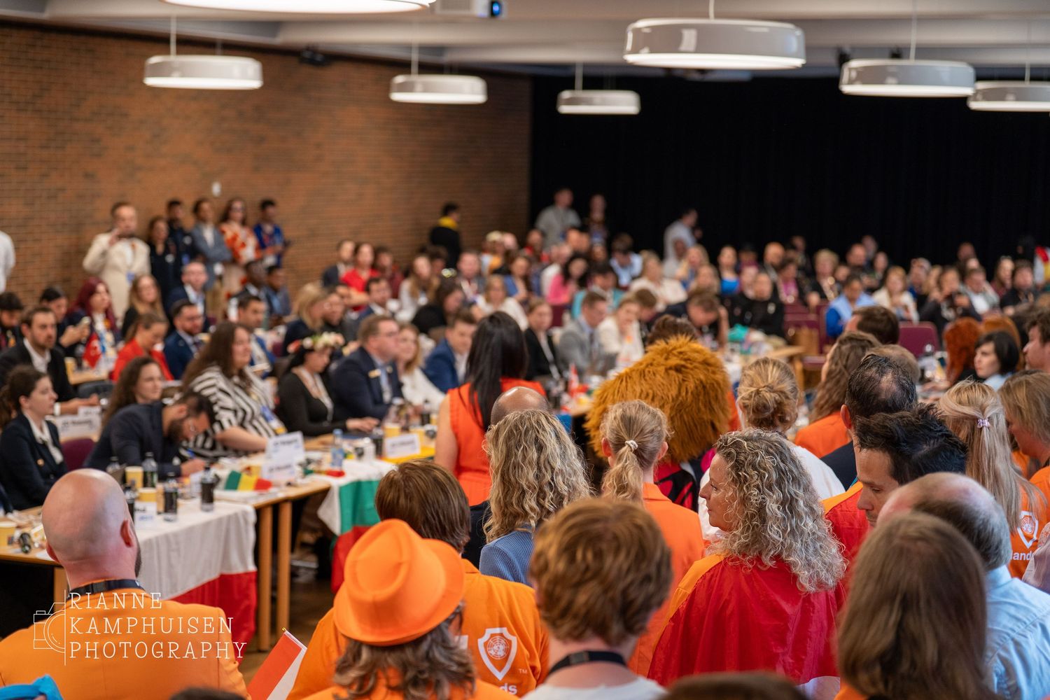 Drukke zaal vol deelnemers, deels in oranje gekleed, tijdens een formele bijeenkomst met internationale vertegenwoordiging.