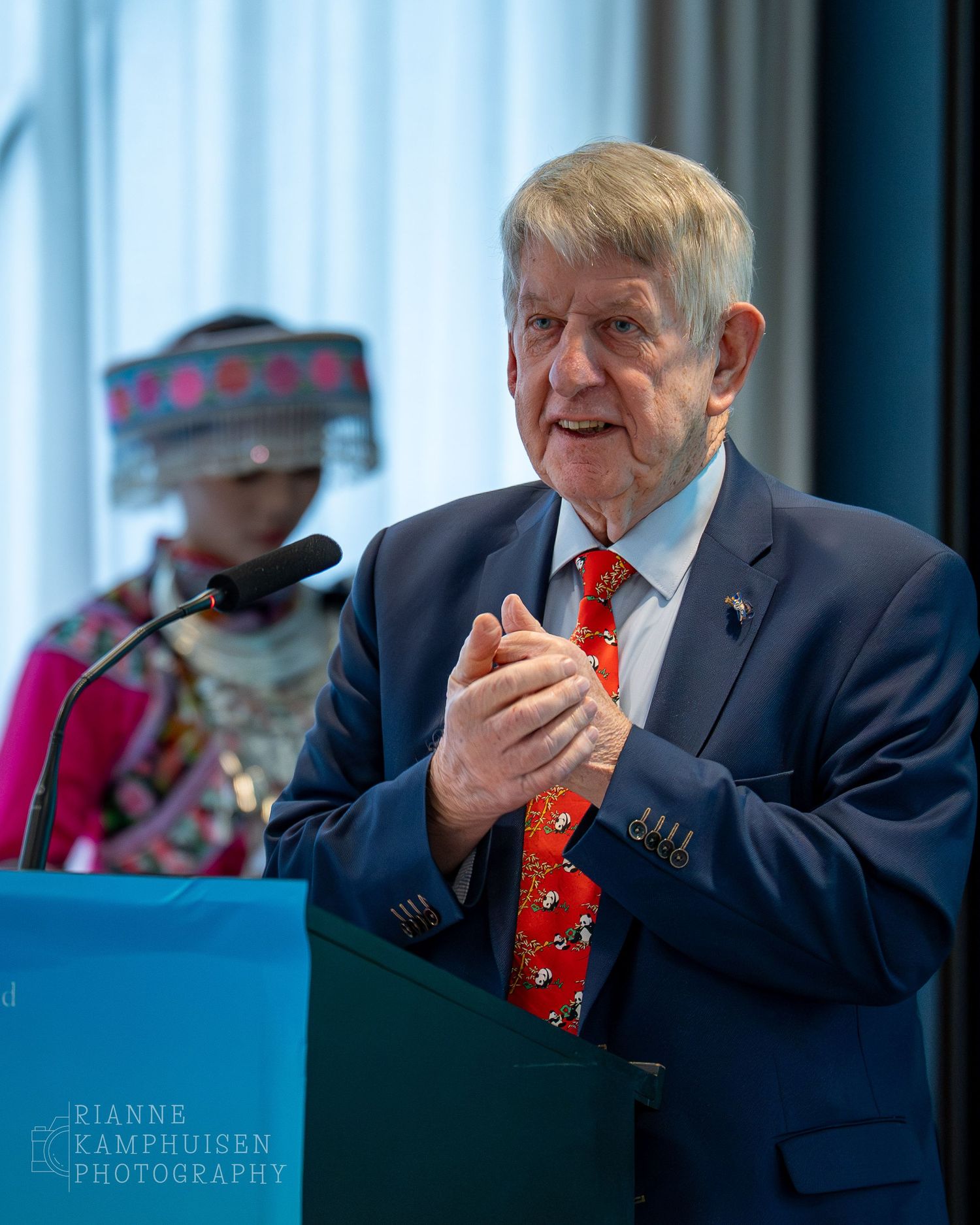 Oudere man spreekt op het podium tijdens de Yibin conferentie, met traditionele Chinese kleding op de achtergrond.