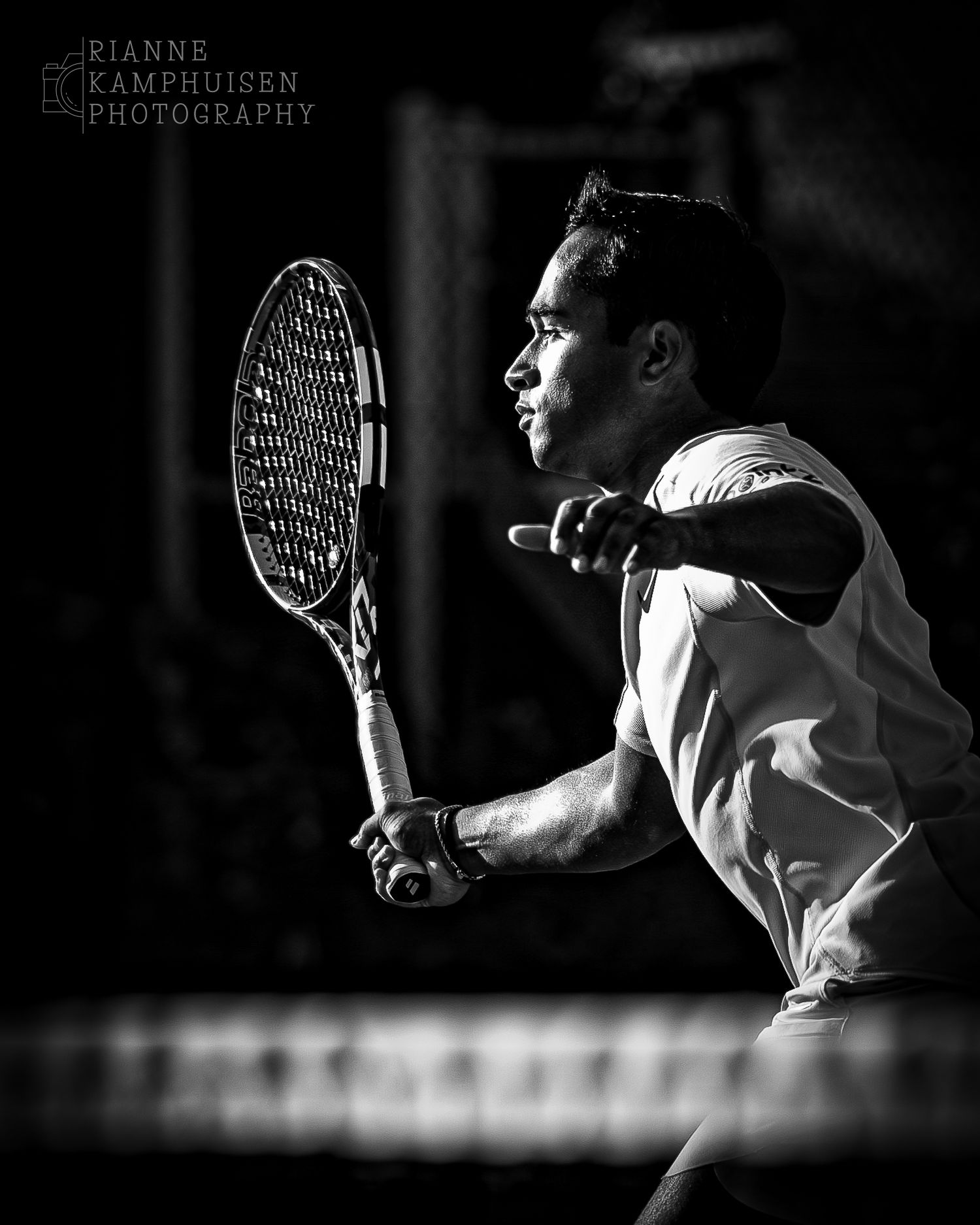 Zwart wit foto van een tennisser die een volley slaat. De zon belicht zijn gezicht