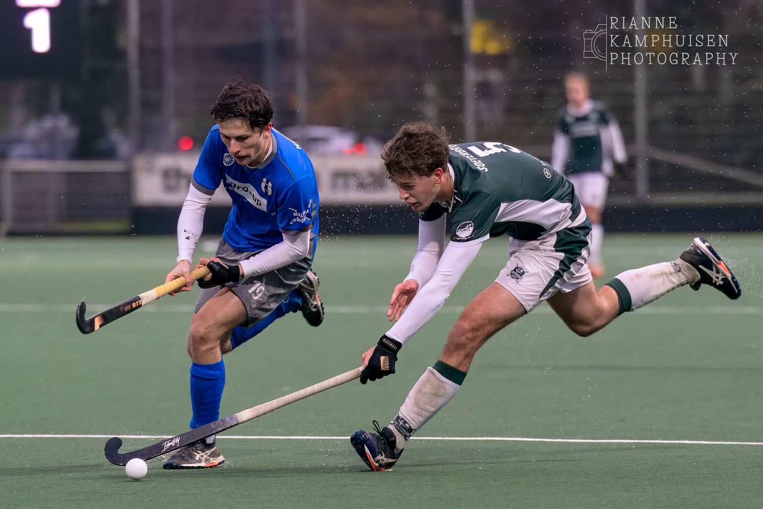 Twee hockeyspelers in duel om de bal op een regenachtig hockeyveld.