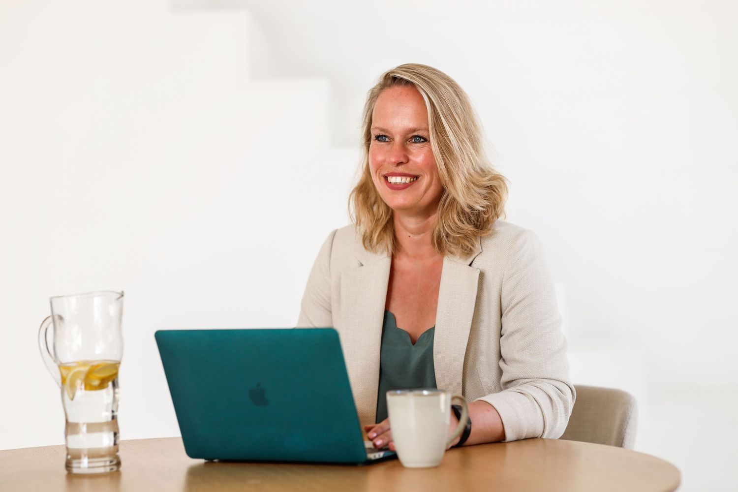 Vrouw werkt glimlachend achter een laptop aan een ronde tafel met een glas water en koffie.
