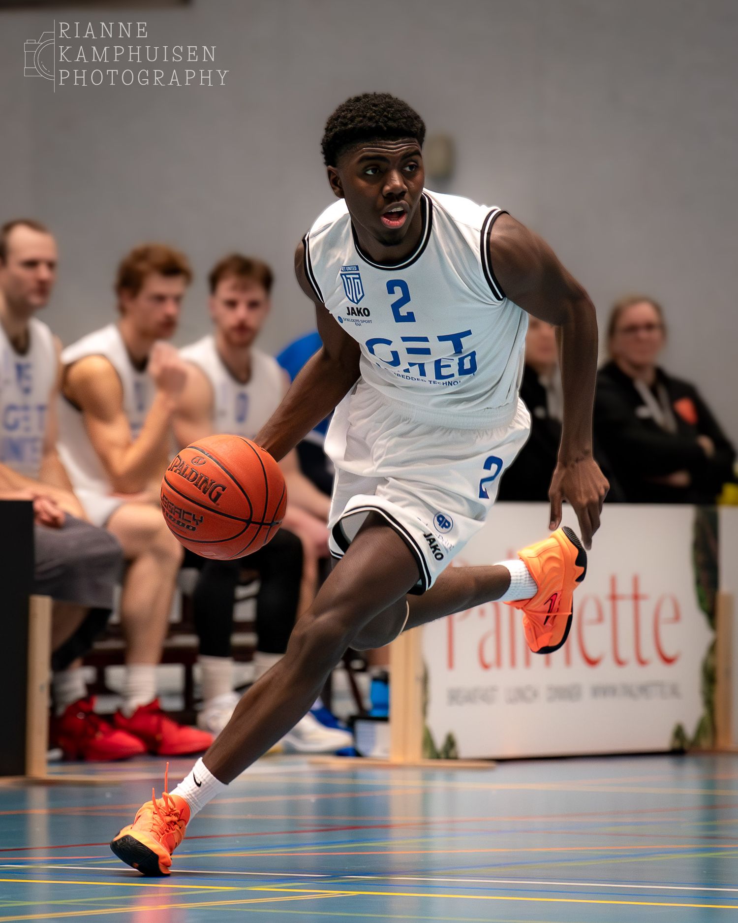 Basketballer in witte tenue die een dribbel maakt om een tegenstander heen, volle focus op rest van sportveld