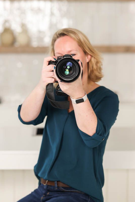 Vrouw met blond haar richt lachend haar camera op de kijker, zittend in een lichte ruimte.