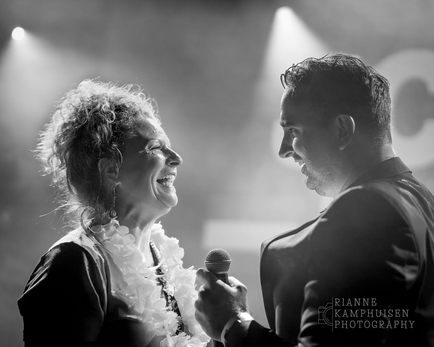 Twee mensen lachen elkaar toe op het podium tijdens een optreden, in sfeervol tegenlicht. Zwart wit foto