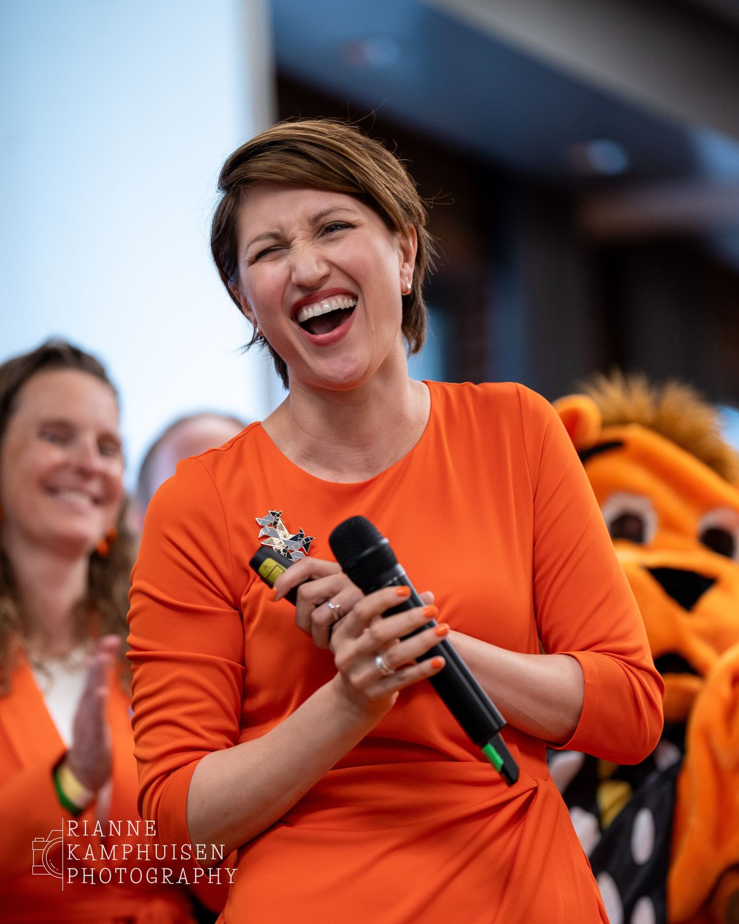 Vrouw in oranje jurk lacht breed met microfoon in de hand, omringd door andere feestelijk geklede mensen en een mascotte op de achtergrond.
