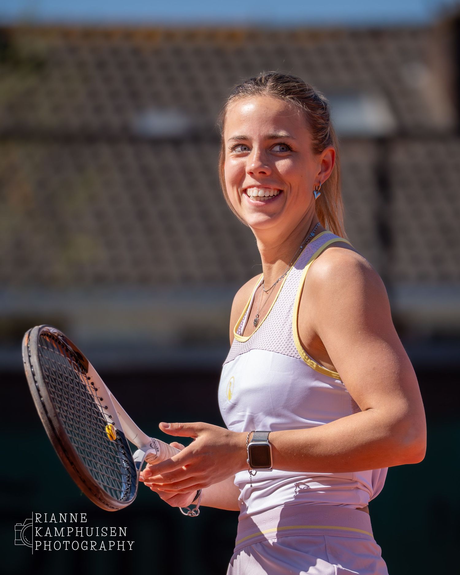 Tennisster in wit sporttenue glimlacht richting de camera tijdens een zonnige speelmoment.