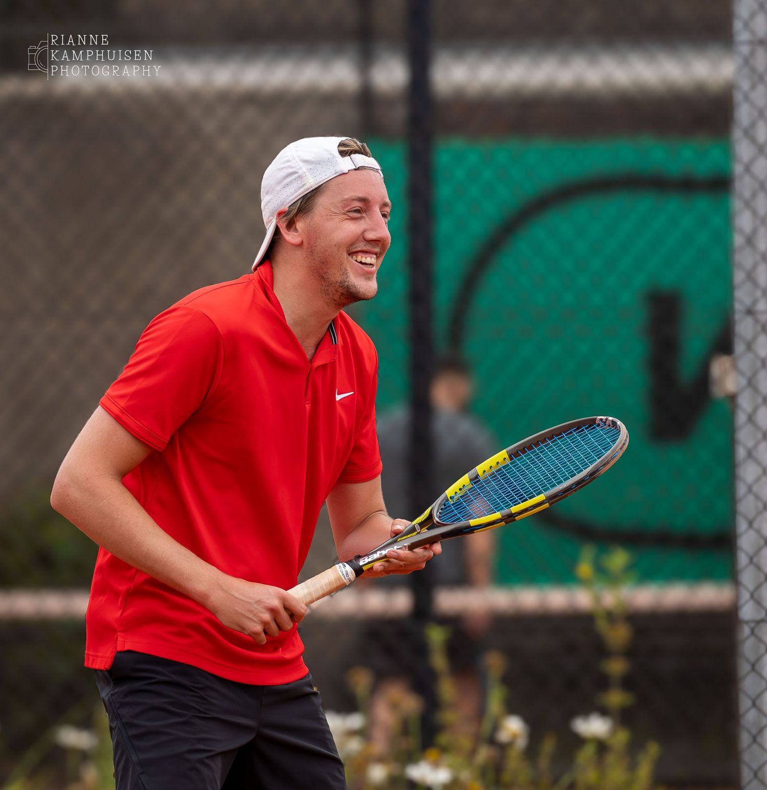 Tennisser in rood shirt en witte pet lacht ontspannen op de baan, racket in de hand.