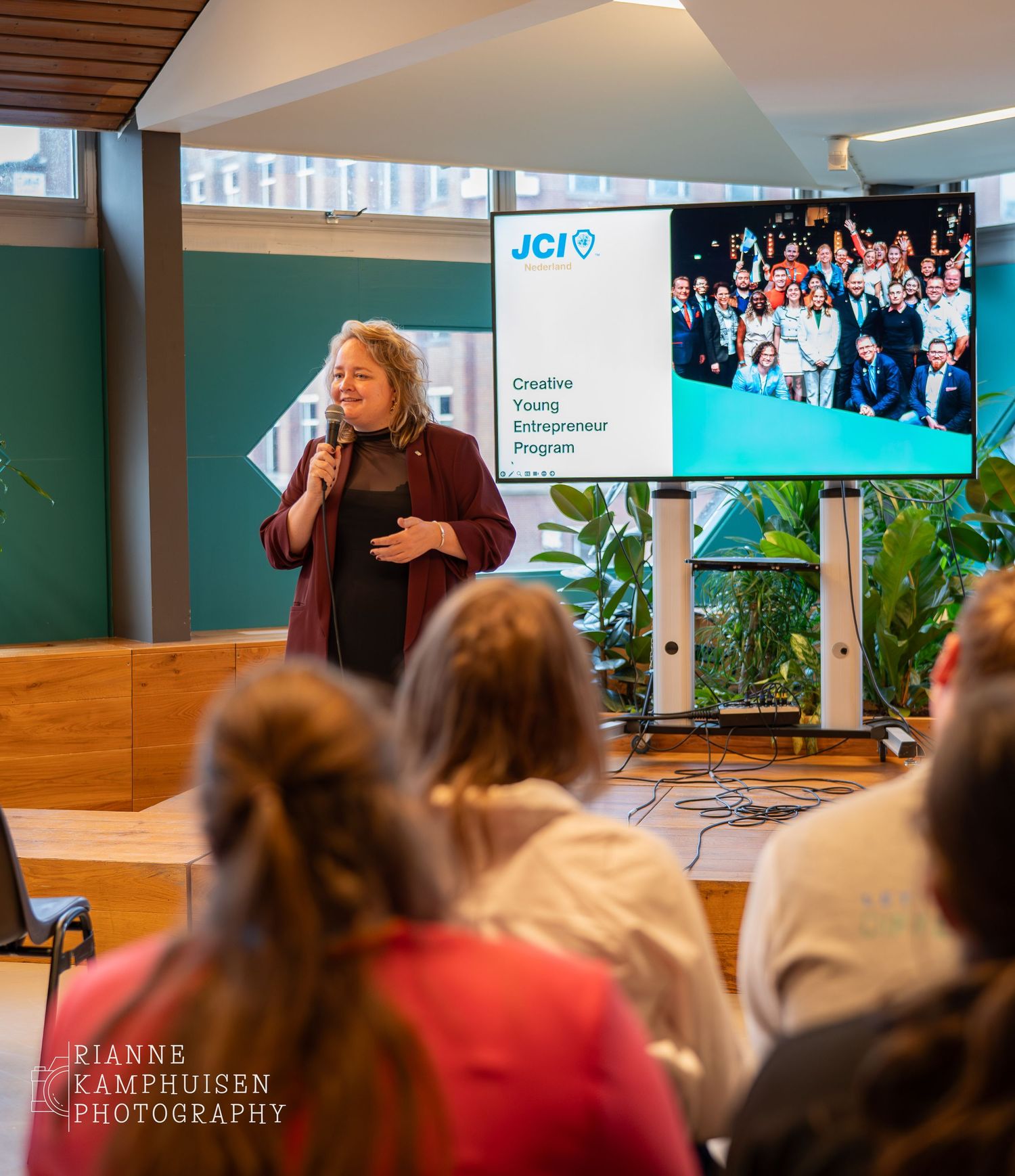 Spreker geeft presentatie voor een groep mensen tijdens een evenement, met een scherm op de achtergrond waarop 'JCI' en een groepsfoto te zi
