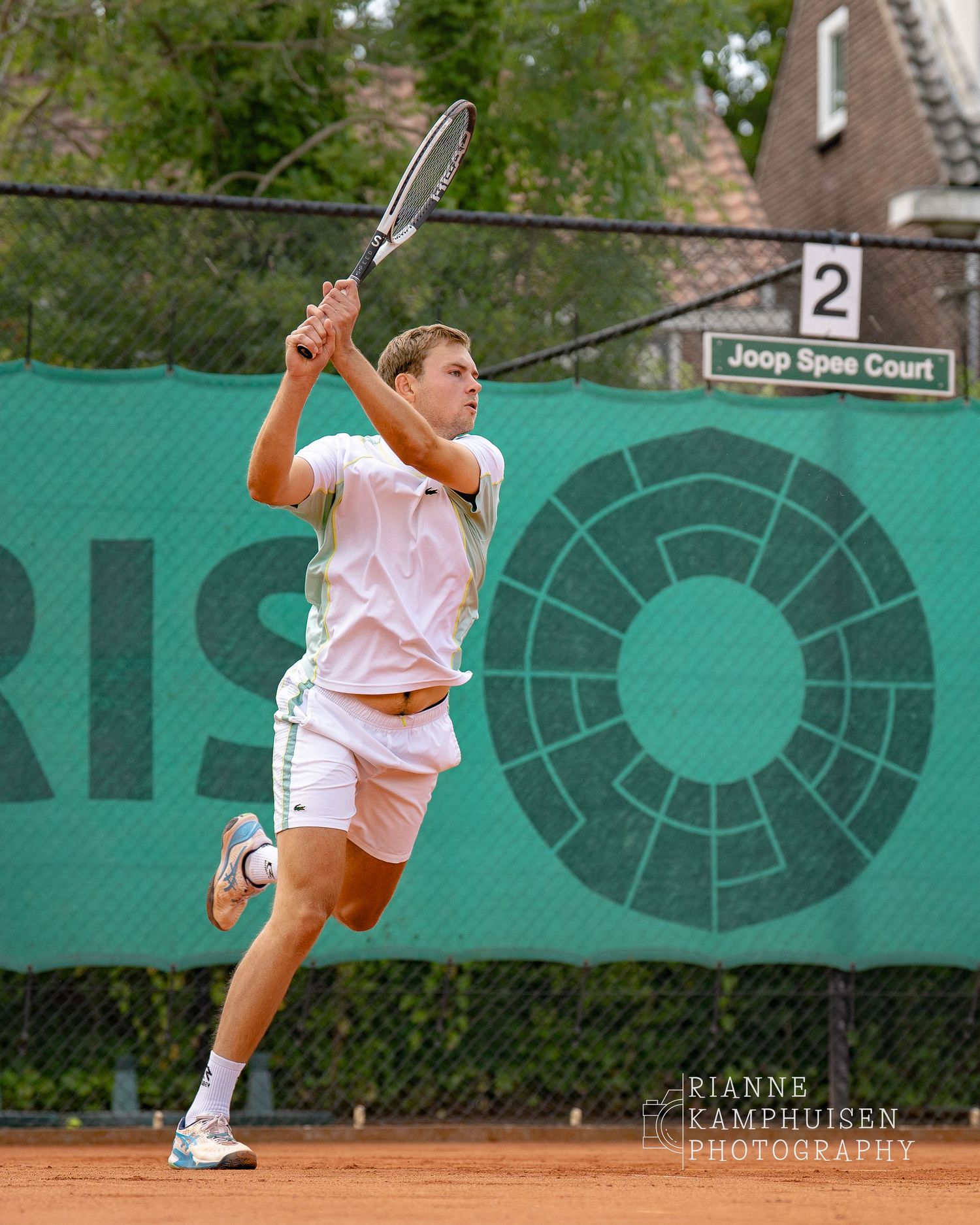 Tennisspeler in witte tenue heeft net een backhand geslagen, hij zit in de uitloop van deze slag.