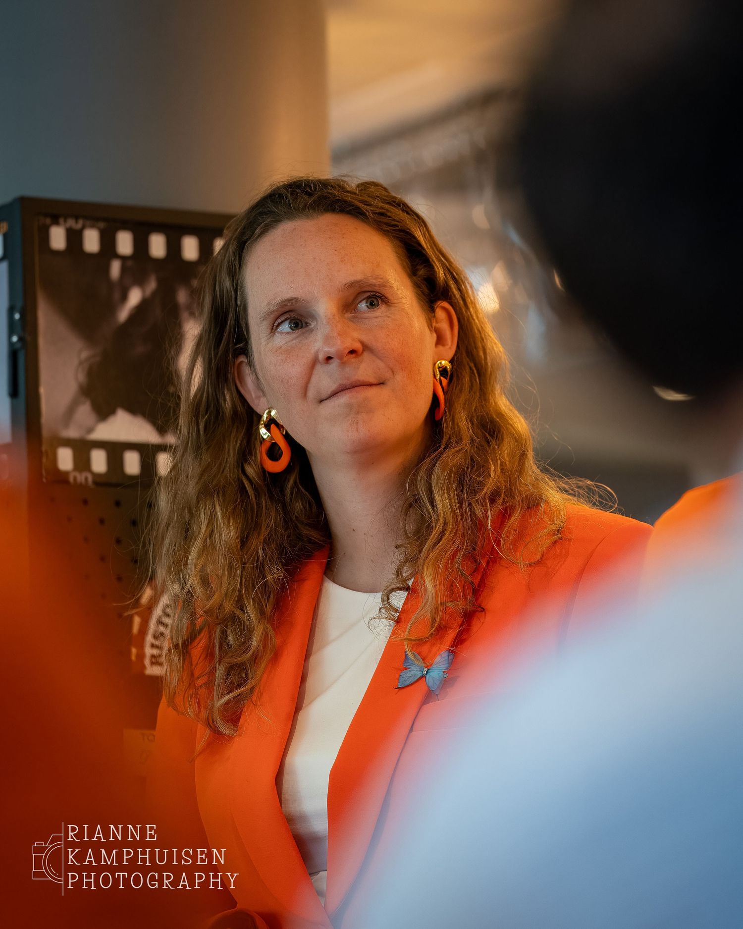 Vrouw in oranje colbert kijkt aandachtig opzij tijdens een bijeenkomst, met warme lichtinval.