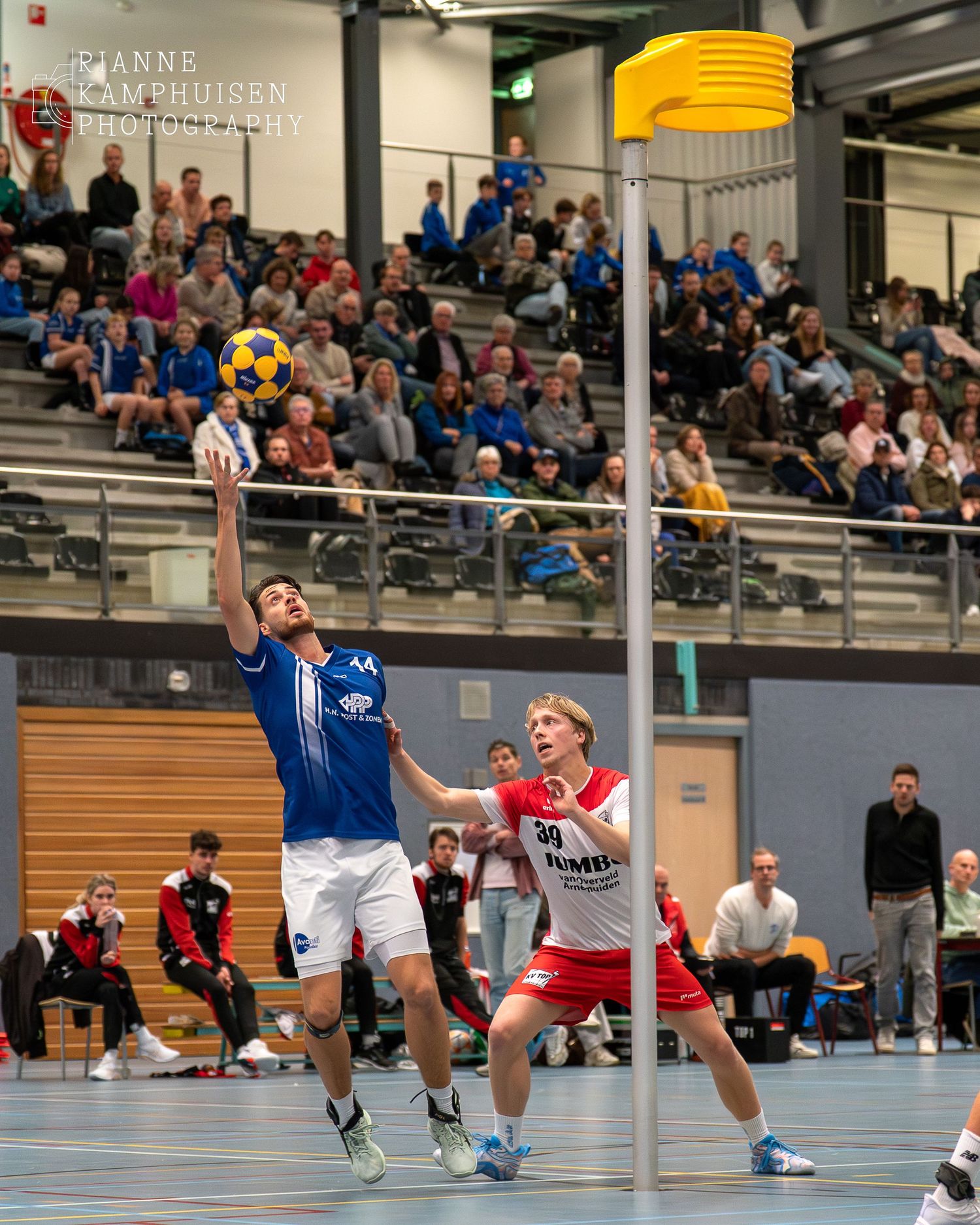 Korfbal speler doet een poging om te scoren, op de achtergrond zie je een volle tribune.