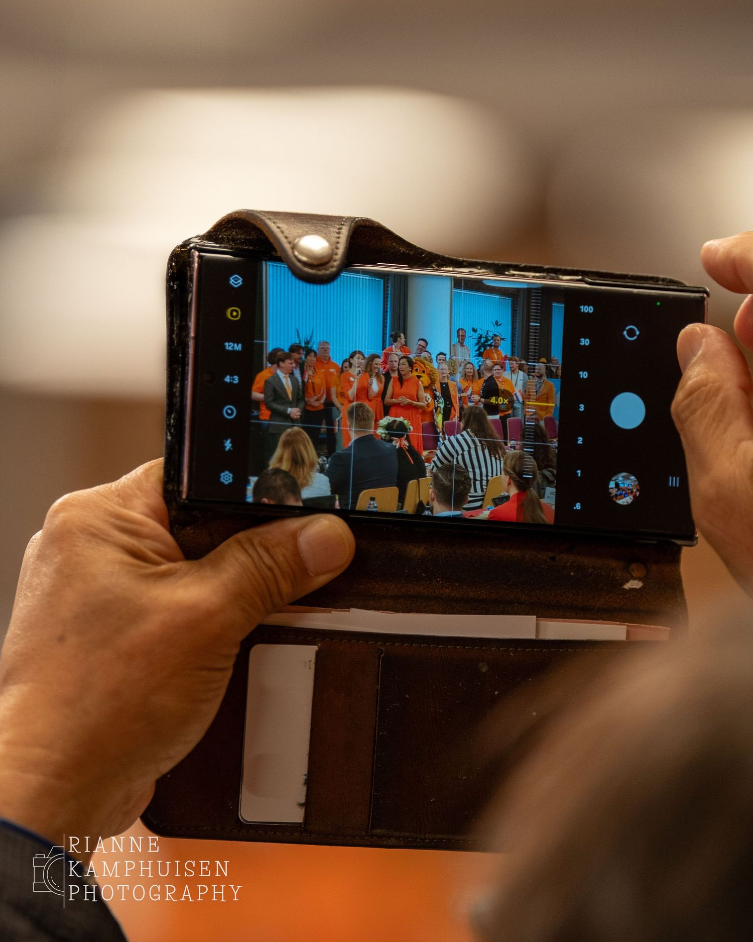 Close-up van een telefoon die een groepsfoto maakt tijdens een evenement, vastgehouden in een leren hoes.