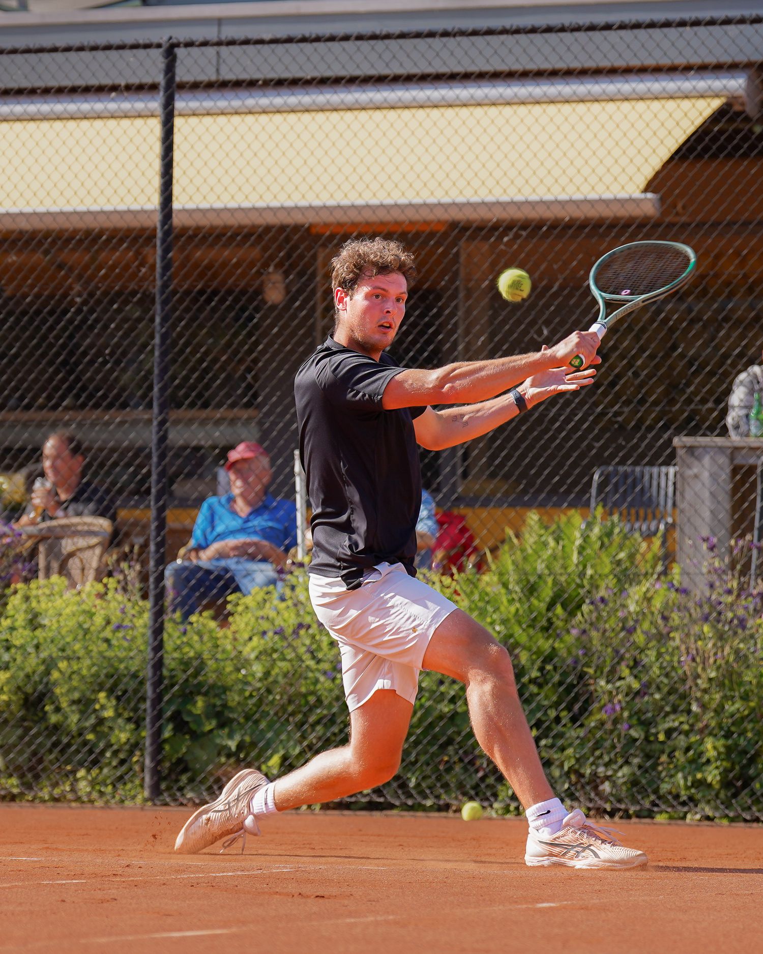 Speler in zwart shirt en witte shorts slaat een backhand terwijl hij zijn lichaam volledig meedraait.