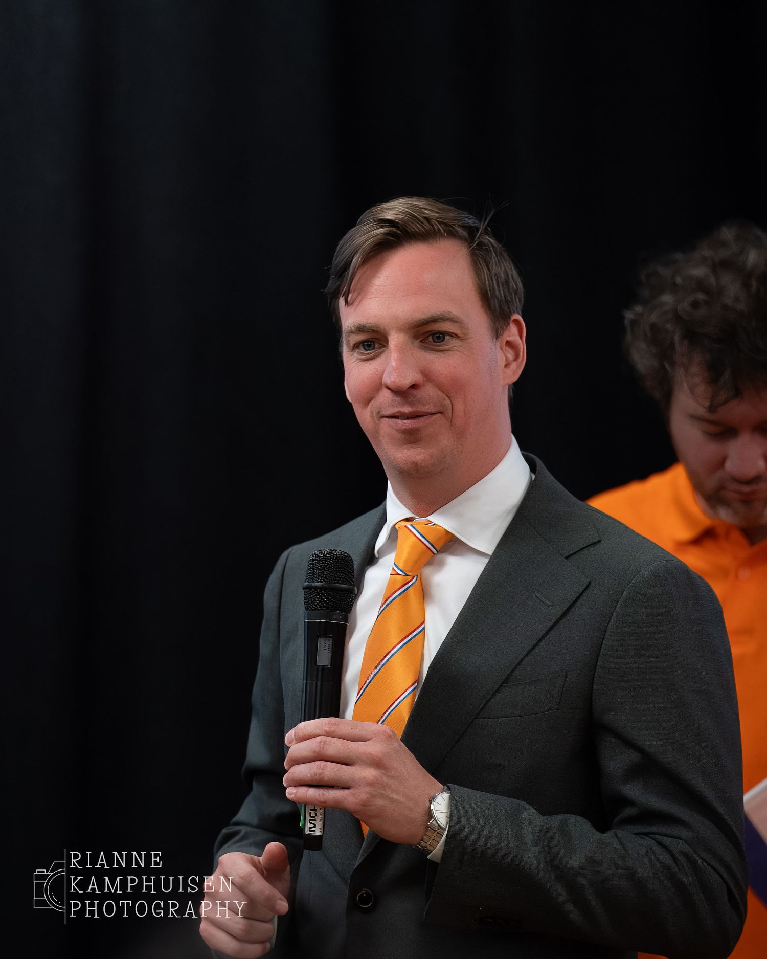 Man in pak met oranje stropdas spreekt met microfoon tijdens een evenement, met een collega in oranje shirt op de achtergrond.