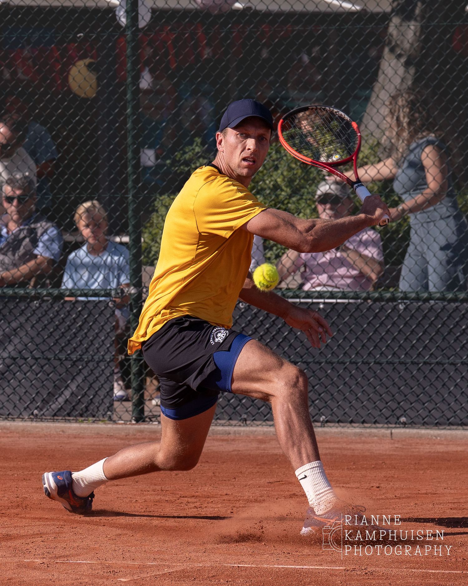 Tennisspeler met geel shirt glijdt naar een bal toe om een slice backhand te slaan tijdens een toernooi in Reeuwijk